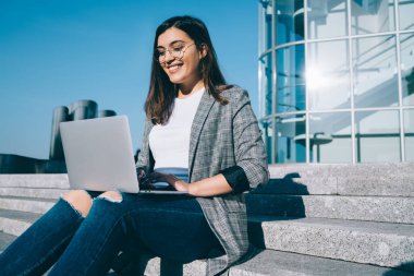 Şirin tebessümlü, klavyeli güzel bir kız öğrenci üniversitedeki derslerden sonra dinlenirken dışarıda merdivenlerde otururken dizüstü bilgisayarında çalışan neşeli mutlu bir kadın.