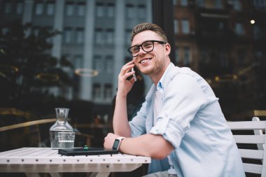 Sıradan gömlekli ve gözlüklü yakışıklı bir genç sokakta masada otururken cep telefonuyla konuşuyor.