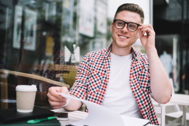 Gözlüklü, mutlu, beyaz bir adam elinde not defteriyle masada oturuyor kahve içiyor ve gülümsüyor.