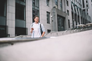 Sırt çantalı genç adam elinde cep telefonuyla caddede dikiliyor ve gülümsüyor.