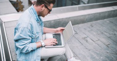 Sokak çitlerinde oturup çalışırken beyaz ekranlı dizüstü bilgisayar kullanan havalı ve yakışıklı bir adamın yan görüntüsü.