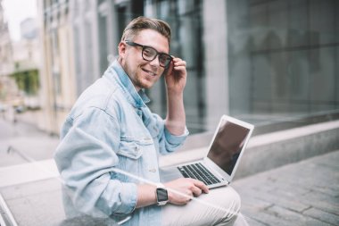 Gözlüklü, kot gömlekli, dizüstü bilgisayarlı ve cam çitin arkasındaki kameraya gülümseyen yakışıklı modern adamın yan görüntüsü.