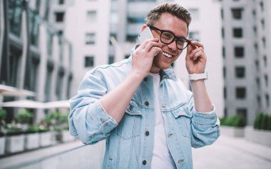 Modaya uygun kot ceketli ve gözlüklü yakışıklı adam mutlu bir şekilde gülümsüyor ve akıllı telefondan sohbet ediyor. Bulanık arka planda duruyor.