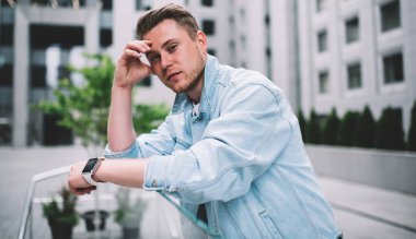 Modern kot ceket giyen, camdan çitlere yaslanan ve sokaktaki kameraya bakan binlerce yıllık beyaz adamın yan görüntüsü.