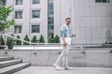 Binlerce yıllık kot ceketli bir adamın dizüstü bilgisayar taşıması ve sokakta yürüyen ve kameraya gülümseyen bir fincan taze kahve içmesi.