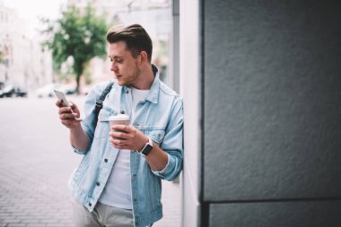 Kot ceketli genç adam elinde bir fincan taze kahve tutarken ve sokakta duvara yaslanırken akıllı telefonları karıştırırken.