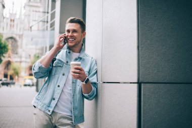Gündelik giysiler içindeki genç adam cep telefonuyla konuşuyor ve kahve içerken gülüyor ve sokakta dikiliyor.