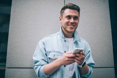 Gri sokak duvarına yaslanıp gülümserken şık kot ceketli, akıllı telefon tutan ve gülümseyen yakışıklı adamdan.