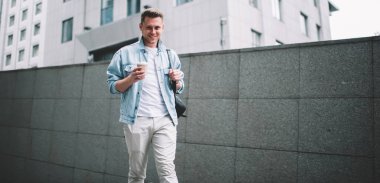 Çağdaş caddede yürürken ve kameraya gülümserken sırt çantasıyla bir fincan taze kahve içen yakışıklı genç adam.