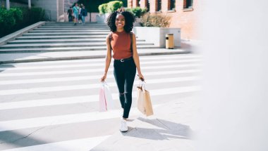 Başarılı esmer kadın portresi. Günlük kıyafetlerle süslenmiş. Alışverişten heyecanlanmış. Elinde kağıt torbalarla sokakta yürüyen pozitif bir kadın.