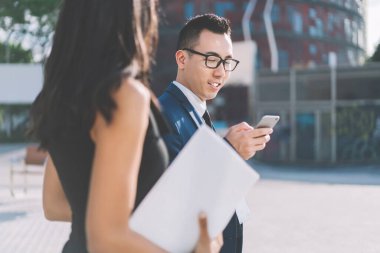 Gözlüklü genç bir Asyalı kadın meslektaşıyla sokakta yürürken akıllı telefon kullanıyor ve konferansta güneş ışığında sohbet ediyor.
