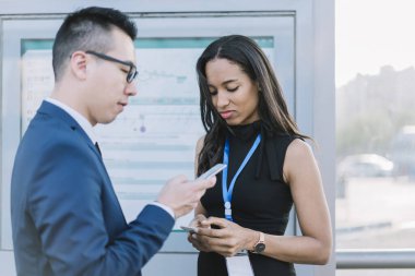 Zarif etnik kadın ve resmi üniformalı Asyalı adam demiryolu istasyonunda akıllı telefonlar kullanarak birlikte duruyorlar. 