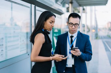 Çok ırklı genç bir kadın ve kimlik kartı olan bir adam şehir tren istasyonunun konferansında yan yana dururken cep telefonu kullanıyor.