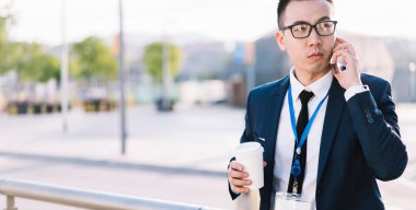 Son moda takım elbiseli ve rozetli genç bir Asyalı elinde kahve fincanı tutarken sokakta çitin başında dikilip telefonla konuşuyor.