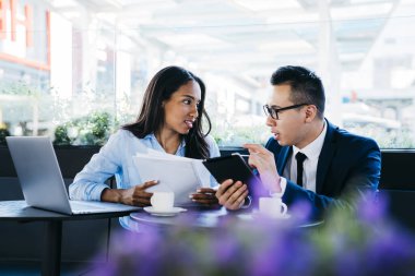 Genç Asyalı erkek ve Afrikalı Amerikalı kadın, kafede oturmuş, profesyonel bir toplantı yaparken, belgeleri tartışıyor ve tablet kullanıyor.
