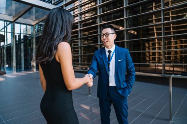 Takım elbiseli ve gazlı mutlu Asyalı bir adam sokakta durup konferansta birbiriyle buluşan genç bayan meslektaşının elini sıkıyor.