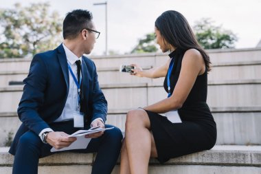 Moda çeşitliliği olan kadın ve erkek sokakta beton tribün üzerine oturmuş cep telefonu kullanarak selfie çekiyorlar.