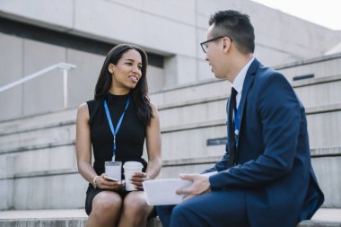 Genç, şık, Afro-Amerikan kadın, erkek iş arkadaşı, kimlik kartı takmış, kahve içiyor ve sokak tribününde oturmuş sohbet ediyor.