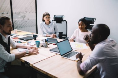 Yeni tasarım projesinde işbirliği yapan çok kültürlü erkek ve kadın meslektaşlar modern ofis içlerinde brifing toplantısı yapıyorlar, profesyonel iş adamlarından oluşan bir grup masada fikirleri tartışıyor.