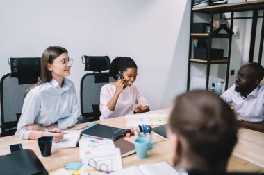 Mutlu Afro-Amerikan bayan çalışanları arkadaşlarıyla akıllı telefon sohbetinin keyfini çıkarırken çeşitli meslektaşlar toplantı masasında oturuyor ve içeride beyin fırtınası sırasında proje detaylarını tartışıyorlar.