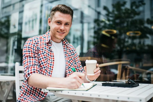 Handsome Millennial Mann Rutete Skjorte Som Holder Kaffekopp Skriver Notisblokk – stockfoto