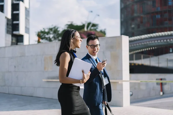 Side view of cheerful modern diverse man and woman in elegant clothes and id badges walking with papers and smartphone on street