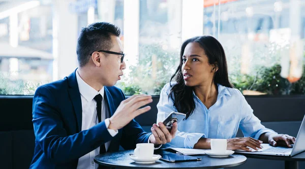Young African American woman with Asian man in formal clothes having business meeting in cafe drinking coffee and using devices while talking
