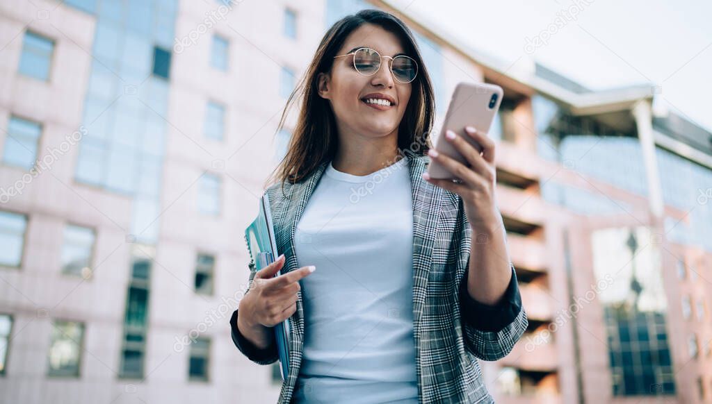 Attractive woman in eyewear for vision correction checking email from colleague during break on urban setting, positive female manager in formal jacket reading funny news on content profile