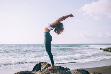 Eğri büğrü kurbağa pozu veren esnek bir kadının doğa ortamında yoga yapması, sağlık motivasyonu ve okyanus sahillerinde enerji uyumu meditasyonu kavramının tadını çıkarması.