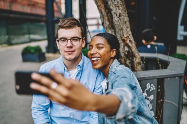 Neşeli afro-amerikan genç kadın akıllı telefonun önünde beyaz tenli hippi adamla birlikte poz vererek internet sitesinde otururken fotoğraf çekiyor. Çeşitli arkadaşlar selfie çekiyor.