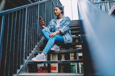 Afrikalı Amerikalı genç kadın blogcu elinde akıllı telefon cihazıyla 4G internet bağlantısı kullanarak takipçilerle mesajlaşıyor. Kot pantolonlu koyu tenli hippi kız cep telefonundan haber okuyor.