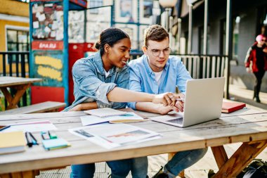 Kot giymiş pozitif çok kültürlü öğrenciler gülümseyip özel ders alıyorlar ve açık havada dizüstü bilgisayarda kablosuz internet kullanıyorlar. Neşeli çeşitli tasarımcılar yayın okuyor.
