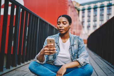 Kot ceketli düşünceli Afro-Amerikalı bir kadın cep telefonundan gelen mesajları okuyor açık havada oturuyor, koyu tenli hippi kız akıllı telefonunu internete bağlayıp blog yazıyor.
