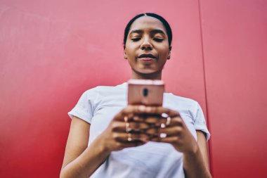 Ciddi koyu tenli kadın blogcu cep telefonu kullanıyor ve dışarıda sohbet ve iletişim kurmak için 4G bağlantısı kullanıyor. Kırmızı duvar arkasında duran akıllı telefonu kullanan düşünceli Afrikalı Amerikalı kadın.