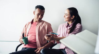 Elinde akıllı telefon cihazıyla sosyal ağlardaki komik mesajlardan gülümseyen ve yanında oturan erkek meslektaşına yayınını gösteren neşeli hippi kız akıllı telefon kullanan olumlu erkek ve kadın.