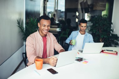 Mutlu Afrikalı Amerikalı erkek yazılım geliştirici modern laptop cihazıyla ofis masasında oturuyor ve komik video, teknoloji ve iletişim konsepti ile e-posta yoluyla gülümsüyor.