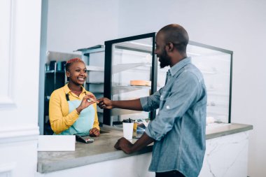 Yakışıklı Afro-Amerikalı adam gülümsüyor ve modern kafeden yemek alırken dost canlısı bayan baristaya kredi kartı veriyor.