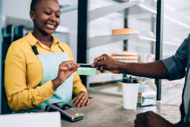 Yetişkin Afro-Amerikan barista gülümseyen ve ekin siyahından kredi kartı alan modern kafede tezgahın arkasında durup yiyecek satan kişi.