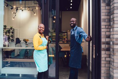 Kafe girişinin yanında durup müşterileri davet ederken önlüklü, gülümseyen ve kameraya bakan Afro-Amerikan erkek ve kadınların yan görüntüsü.