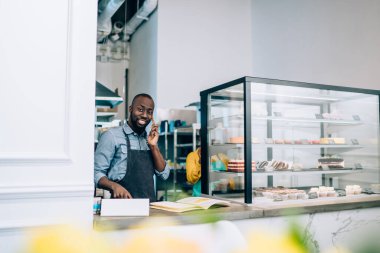 Yakışıklı Afro-Amerikalı adam gülümseyip kameraya bakarken akıllı telefon konuşması yapıyor ve küçük bir kafede tezgahın arkasında duruyor.