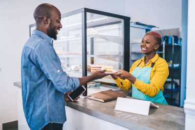 Dost canlısı Afrikalı Amerikalı bayan barmen neşeyle gülümsüyor ve küçük bir pastanede çalışırken siyah erkek müşteriye bir parça taze kek veriyor.