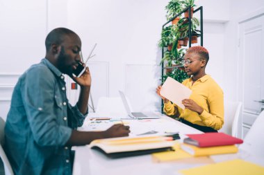 Afro-Amerikalı bir adam telefonda konuşurken, siyahi bir kadın kafe masasında birlikte otururken gazetelerle çalışıyor.