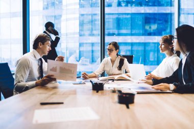Modern ofis yönetim kurulundaki masada istatistiksel dokümanlar tartışılırken, takım halinde çalışan çağdaş kadın ve erkek grupları 