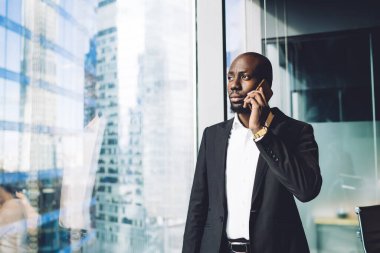 Ofisin penceresinin yanında duran, cep telefonuyla konuşurken şehir manzarasına bakan takım elbiseli yakışıklı, Afro-Amerikan bir adam.