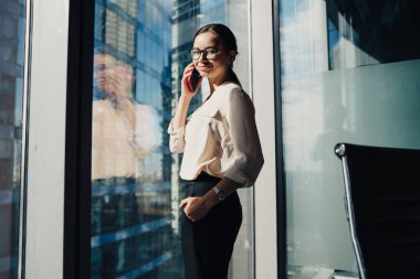 Gözlüklü, neşeli, zarif bir kadının güneşli bir ofiste kameraya gülümserken telefon etmesi.