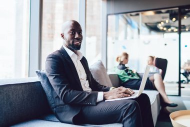 Ofis koridorundaki koltukta oturan şık, yakışıklı, Afro-Amerikan bir adam dizüstü bilgisayarla kameraya gülümsüyor.