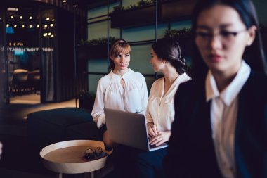 Resmi giyinmiş modern kadınlar ofis koridorunda kanepede dizüstü bilgisayar kullanarak otururlar ve çalışırken profesyonel tartışmalar yaparlar. 