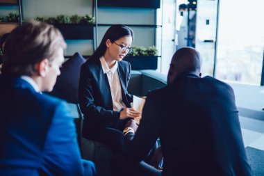 Bardaklı genç Asyalı kadın ofis koridorunda çeşitli seçkin iş adamlarıyla oturuyor belgeleri paylaşıyor ve takım çalışması yapıyor. 