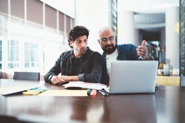 Modern ofisteki kırtasiye malzemeleriyle masada otururken grup halinde proje üzerinde çalışan ve dizüstü bilgisayarını paylaşan modaya uyan genç erkekler.