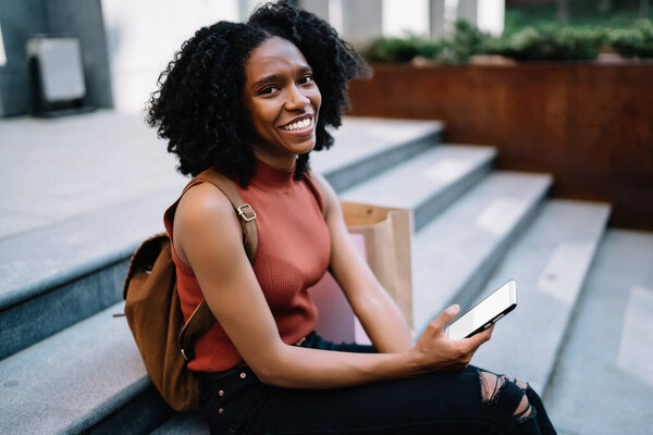 Portrait of happy female teenager with modern digital gadget in hands laughing during online communication with followers, cheerful dark skinned woman in casual wear taking rest on urban setting